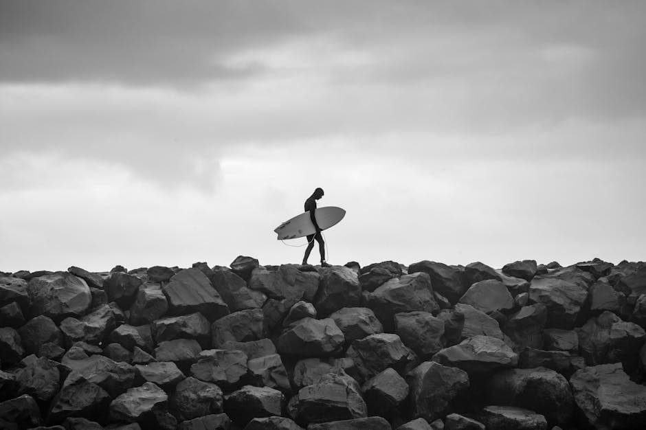 A black and white photo of a surfer walking on rocks